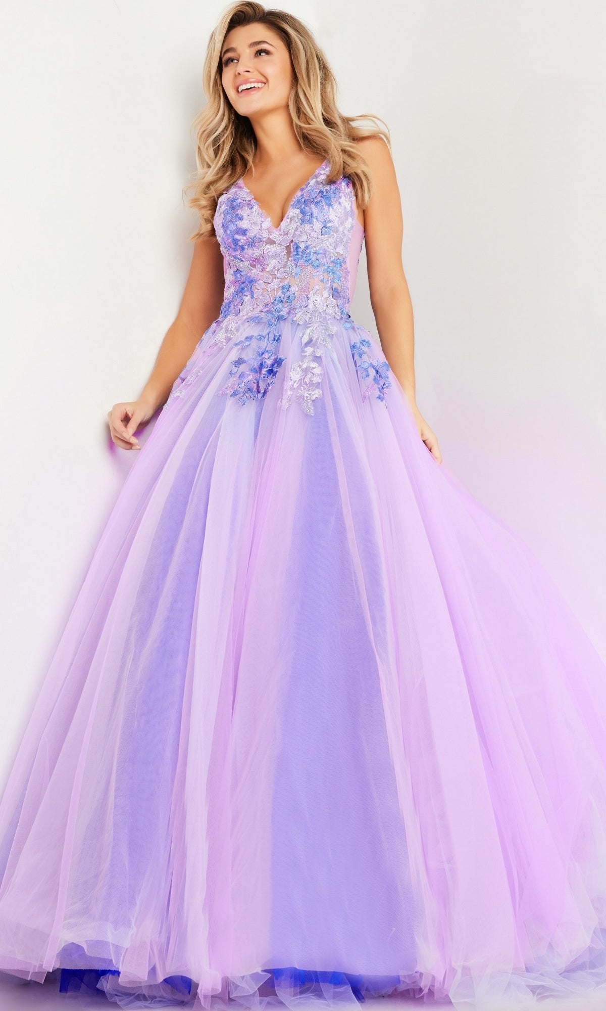 A woman wears the Jovani Lilac Purple Long Prom Ball Gown 24602, a floor-length dress with floral appliqués and an embroidered bodice, standing against a light background.
